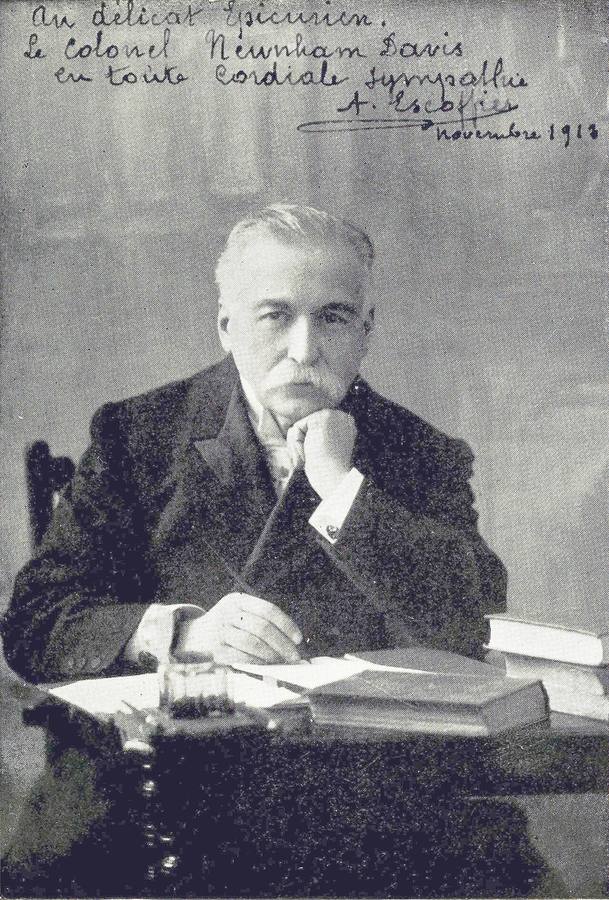 Black and white photo of Auguste Escoffier seated at a desk with books, with a handwritten dedication across the top